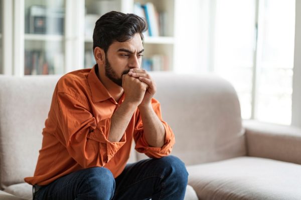 A gentleman in an orange shirt sitting on a couch and contemplating sorrowfully.