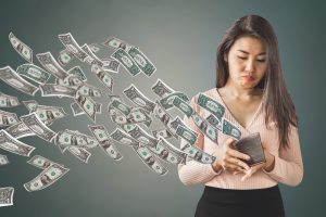 A woman in a peach sweater frowning as she watches several dozen dollar bills fly out of her wallet.